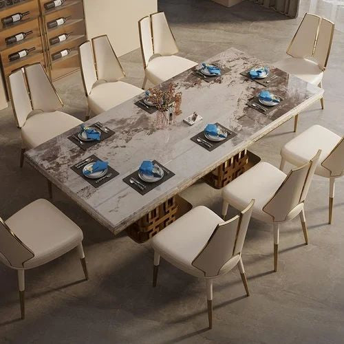 A modern dining room scene with a rectangular marble-patterned dining table surrounded by cream upholstered chairs with gold-toned legs and backs. Blue napkins and black placemats are arranged on the table, which sits on a gray concrete floor in an indoor space with shelving visible in the background.