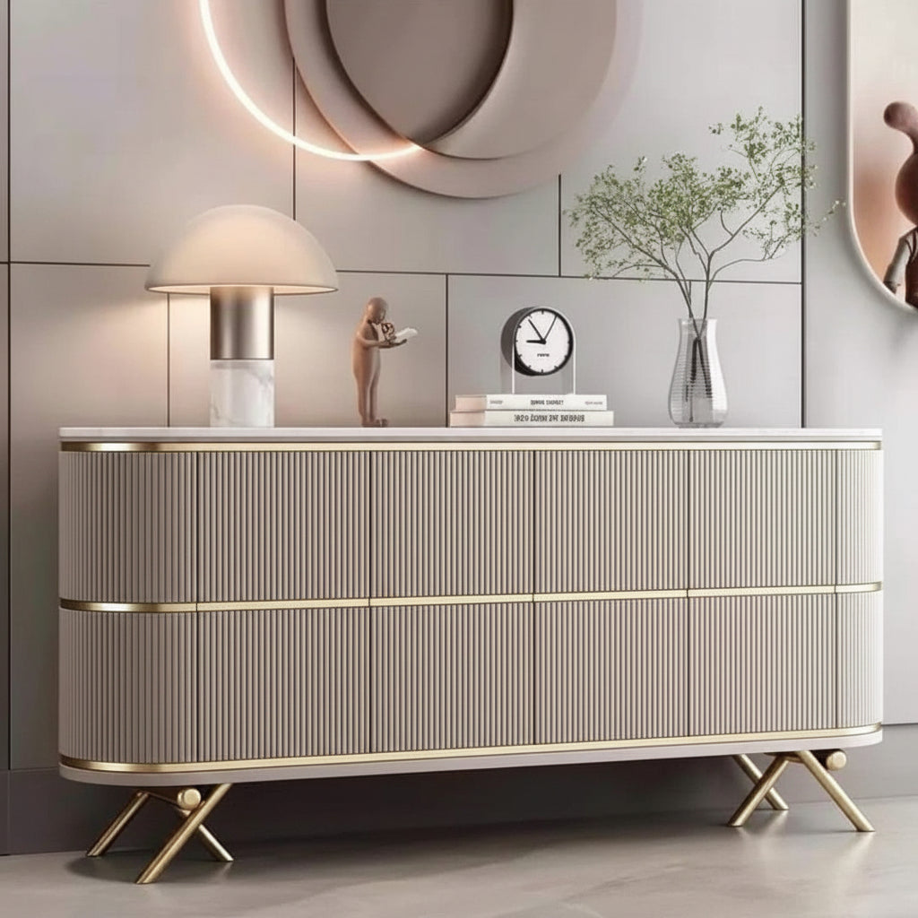 A modern ribbed console cabinet in a warm beige tone with gold trim and gold legs, styled in a contemporary living room setting with a lamp, clock, and decorative vase on top.
