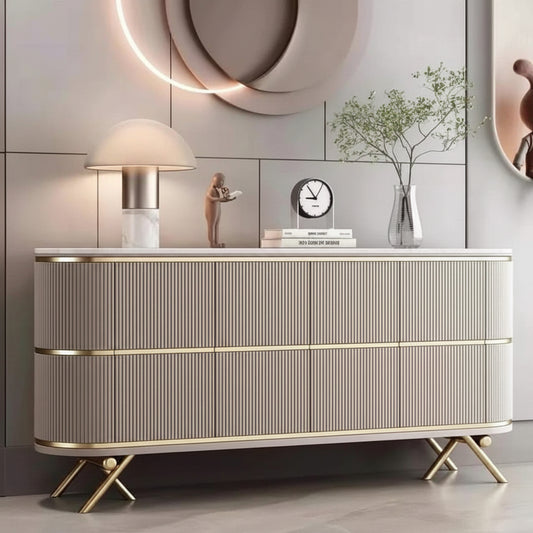 A modern ribbed console cabinet in a warm beige tone with gold trim and gold legs, styled in a contemporary living room setting with a lamp, clock, and decorative vase on top.