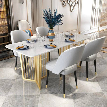 A modern dining table set in a bright, elegant dining room. The table features a white marble-look top with a gold geometric wire base, surrounded by six light gray upholstered dining chairs with black legs and gold accents.