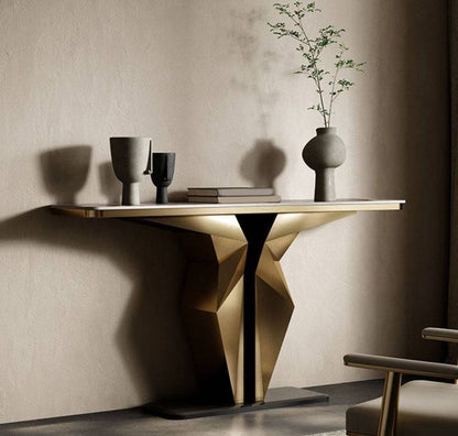 A modern console table with a geometric faceted metallic base and a light wood top, styled in a minimalist room with a textured beige wall, a vase with branches, and a chair partially visible.