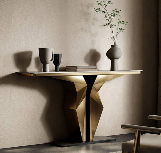 A modern console table with a geometric faceted metallic base and a light wood top, styled in a minimalist room with a textured beige wall, a vase with branches, and a chair partially visible.