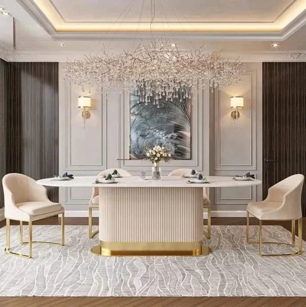 A luxury dining room featuring a modern rectangular dining table with a ribbed cream-white body and gold base, surrounded by four matching cream upholstered dining chairs with gold legs. The room is decorated with a large crystal chandelier, wall sconces, and a patterned area rug.