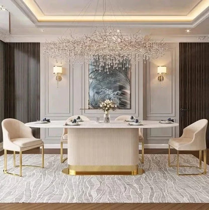 A luxury dining room featuring a modern rectangular dining table with a ribbed cream-white body and gold base, surrounded by four matching cream upholstered dining chairs with gold legs. The room is decorated with a large crystal chandelier, wall sconces, and a patterned area rug.