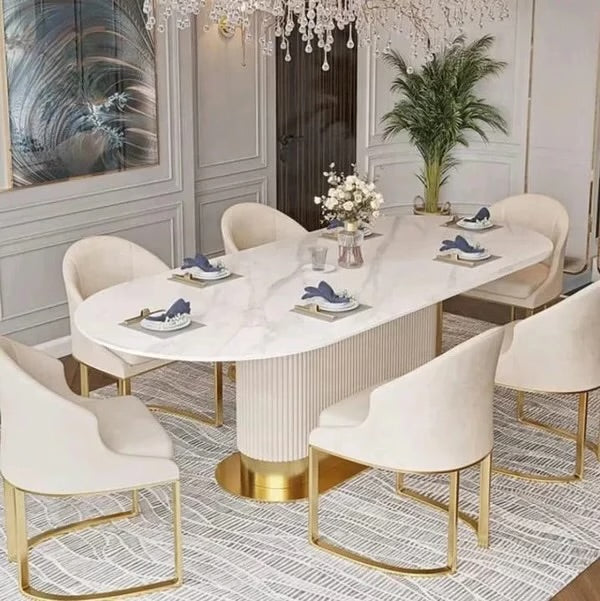 An elegant oval dining table with a white marble-look top and ribbed pedestal base, surrounded by six cream upholstered dining chairs with gold metal legs, set in a luxurious dining room.