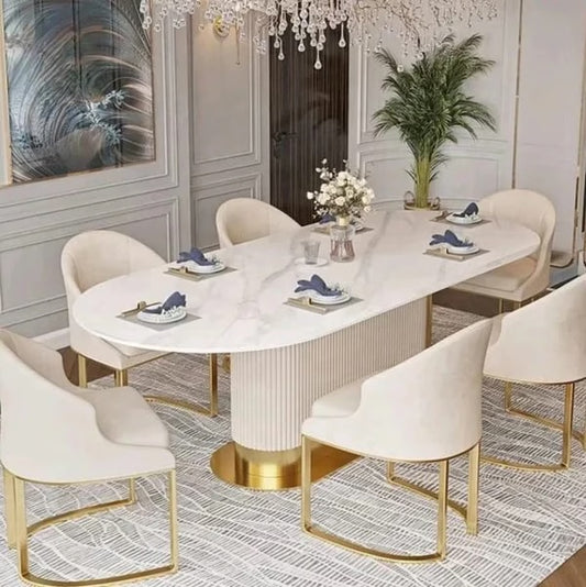 An elegant oval dining table with a white marble-look top and ribbed pedestal base, surrounded by six cream upholstered dining chairs with gold metal legs, set in a luxurious dining room.