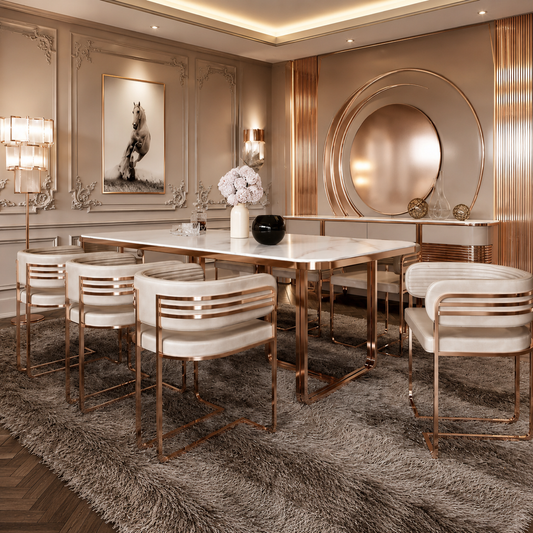 A luxury dining room set featuring a rectangular white marble-top dining table with a polished rose gold metal frame, surrounded by six matching high-back bar stools with white upholstered seats and rose gold metal frames. The set is styled on a plush gray shag rug in an elegant, high-end dining room with gold-toned walls, decorative molding, and a large circular mirror.