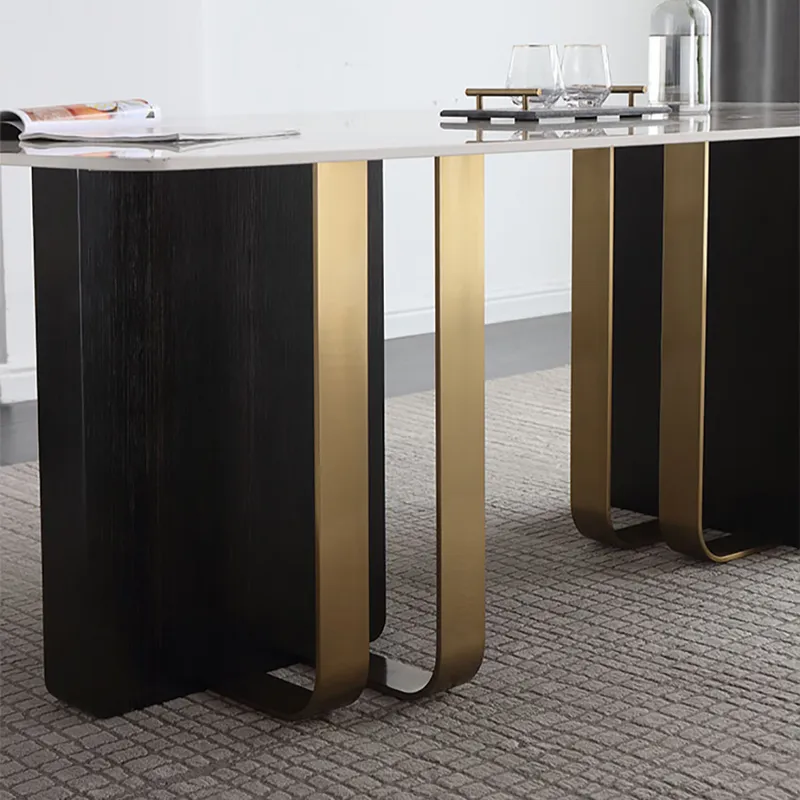 Modern rectangular dining table with a light stone top, black wood-look side panels, and brushed gold U-shaped metal legs, shown in a real interior setting on a textured rug.